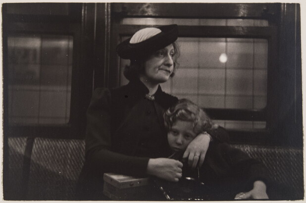A black-and-white photograph of a White woman with her arms around a child; both seated on an upholstered seat with windows behind them.