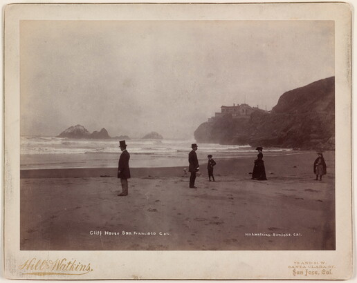 A black-and-white photographs of formally-dressed people scattered along a flat beach and calm shoreline; in the background, a building sits atop a high cliff.