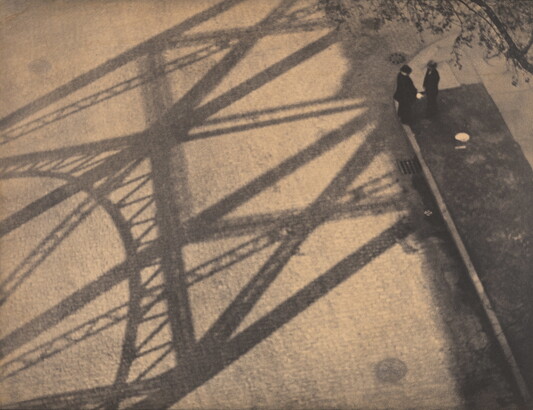 A black-and-white photograph taken from above of two people standing in the shadow of a bridge.