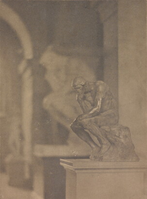 A black-and-white photograph of a small sculpture of a sitting man resting his head on his fist and elbow on one knee in front of a larger sculpture of the same.