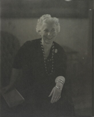 A black-and-white photograph of a smiling White woman with short white hair, seated; one hand holds a book, the other rests on her knee.
