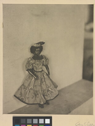 A black-and-white photograph of a dark-colored doll with curly hear wearing a patterned dress and hat posed against a wall.