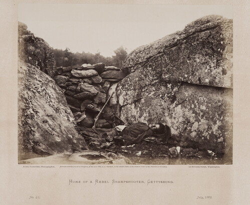 A black-and-white photograph of a dead man lying in a rocky trench next to a rifle.