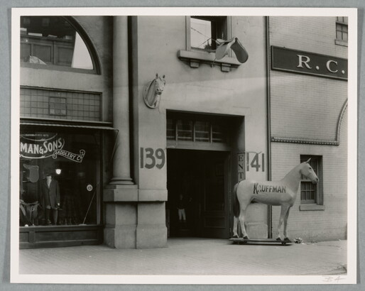 A black-and-white photograph of a storefront with a large statue of a horse, the word "Kauffman" painted on its side, next to the entrance.