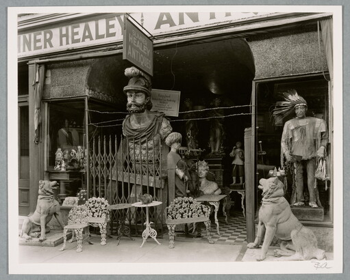 A black-and-white photograph of an overcrowded storefront with a variety of furniture and large statues at the entrance.