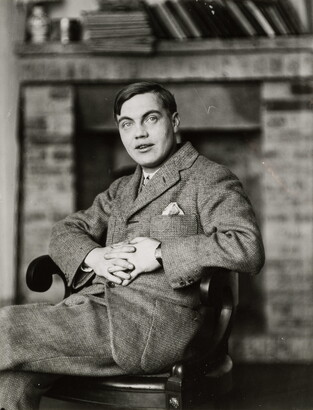 A black-and-white portrait photograph of a White man in a suit sitting in a wood chair with his hands clasped in front of him.