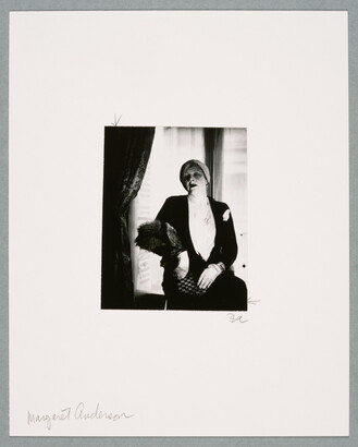 A black-and-white photograph of a White woman, hair wrapped in a turban, seated near a window wearing a black jacket, white gloves, and a fur stole.