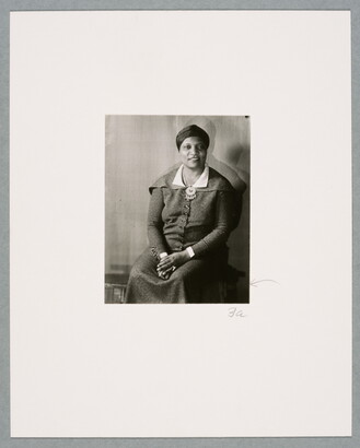 A black-and-white photograph of a seated Black woman, hair wrapped in a turban, wearing a dark suit-dress, hands folded in her lap.