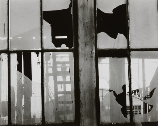 A black-and-white photograph of broken window panes in what looks like a factory building.