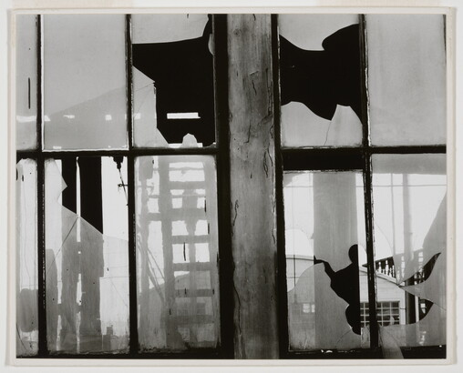 A black-and-white photograph of broken window panes in what looks like a factory building.