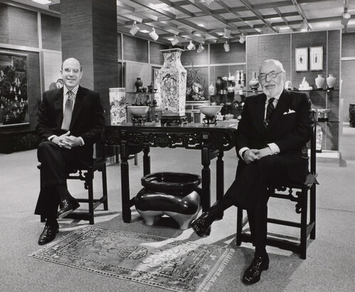 A black-and-white photograph of two smiling White men in suits sitting on either side of a table displaying Asian decor.