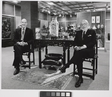 A black-and-white photograph of two smiling White men in suits sitting on either side of a table displaying Asian decor.