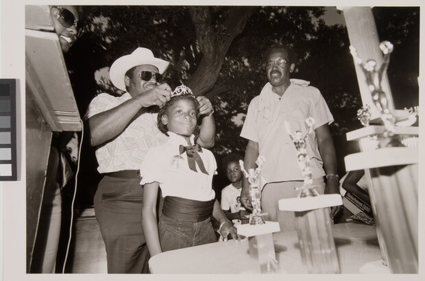 A black-and-white photograph of a young Black girl having a tiara placed on her head as others watch.