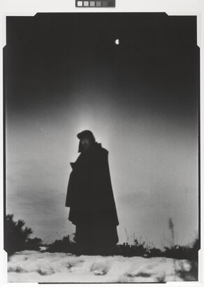 A black-and-white photograph of a heavily cloaked person standing in a frozen landscape with a half-moon behind them.