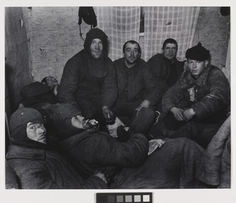 A black-and-white photograph of a group of White men wearing dark, heavy clothing huddled in a small room with plaid curtains over a window.