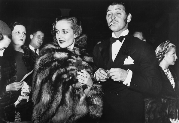 A black-and-white photograph of a White woman wearing a fur coat and a White man with a mustache, smoking a cigarette, wearing a tuxedo.