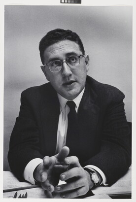 A black-and-white photograph of a White man with short, dark hair in a suit, tie, and glasses leaning forward with his elbows on a desk, gesturing with his hands.
