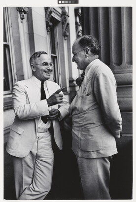 A black-and-white photograph of two White men, both in light-colored suits and glasses, talking to one another and smiling.