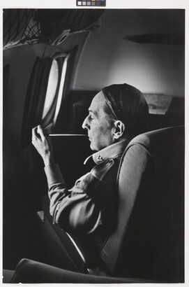 A black-and-white photograph of a White man seated on an airplane smoking a long thin pipe.
