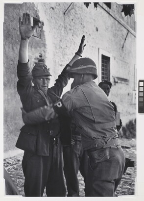 A black-and-white photograph of a White man in military uniform being frisked by a second man in military uniform.