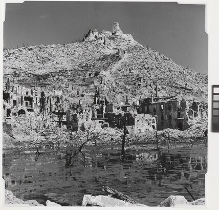 A black-and-white photograph of the ruins of a town at the edge of a body of water and a ruined building on top of a mountain in the background.