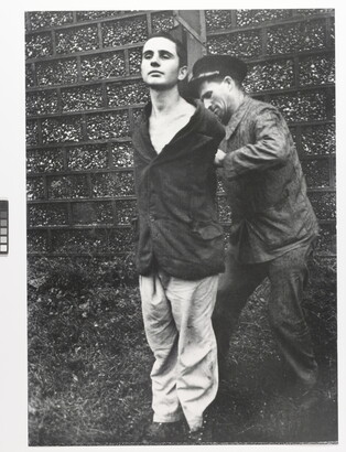A black-and-white photograph of a young, White man standing with his hands behind his back as another White man ties them to a wood pole.