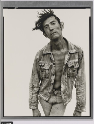 A black-and-white portrait photograph of a White man with dark hair that sticks up, wearing jeans and a jean jacket, and crossing his legs while bending forward.