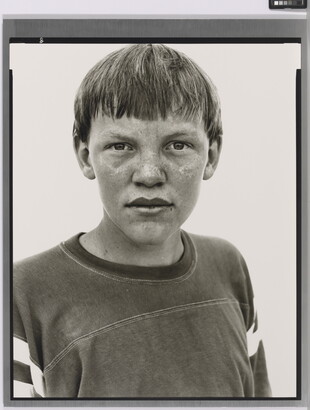 A black-and-white portrait photograph of a White teenage boy with a bowl haircut and freckles across his nose and cheeks, wearing an athletic t-shirt.