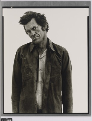 A black-and-white portrait photograph of a White man standing with his head cocked, frowning, and wearing two soiled shirts.