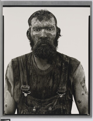 A black-and-white portrait photograph of a bearded man covered in mud and dirt, wearing dirty overalls, and looking straight at the camera.