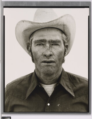 A black-and-white portrait photograph of a White man wearing a cowboy hat and collared shirt, with stubble on his face and chapped lips.