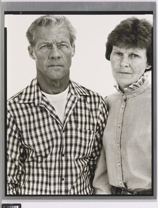 A black-and-white portrait photograph of a middle-aged White man wearing a checked button-down shirt and a middle-aged White woman in a striped high-collar blouse standing shoulder to shoulder.