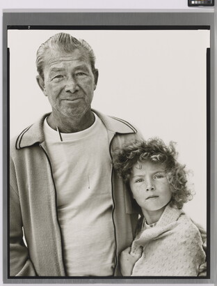 A black-and-white portrait photograph of an older White man in a t-shirt and cardigan with his arm around a young White girl with curly hair and a blanket wrapped around her shoulders.