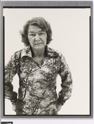 A black-and-white portrait photograph of an older White woman with wavy hair wearing a patterned polo shirt, hands hooked into her back pants pockets.