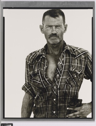 A black-and-white portrait photograph of a middle-aged White man with scruffy facial hair wearing a plaid button-down shirt standing with one hand on his hip.