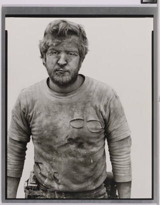 A black-and-white portrait photograph of a White man with messy hair and a beard wearing a dirty, ripped shirt.