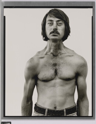 A black-and-white portrait photograph of a shirtless White man, with dark hair and a mustache, and thin but with muscled shoulders and arms.