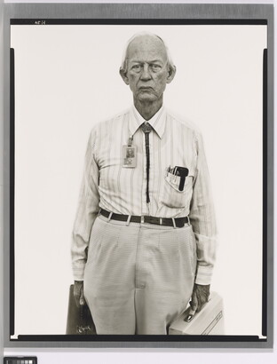 A black-and-white portrait photograph of an older, mostly bald White man wearing a collared shirt, a bolo tie, and work badge attached to his collar, holding two briefcases.