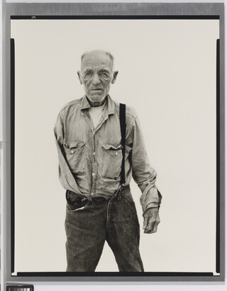 A black-and-white portrait photograph of an older, balding White man with one arm wearing a button-down shirt and jeans held up with one suspender.