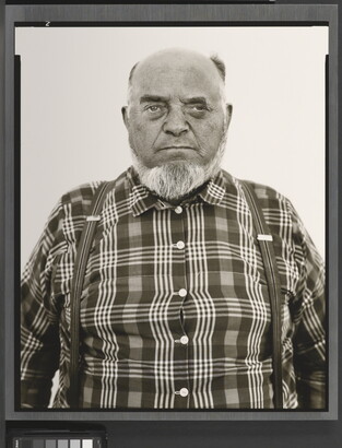A black-and-white portrait photograph of an older, balding White man with a gray beard wearing a plaid button-down shirt and suspenders.