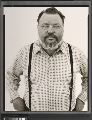 A black-and-white portrait photograph of a middle-aged White man with a receding hairline and salt-and-pepper beard wearing a checked button-down shirt and suspenders.