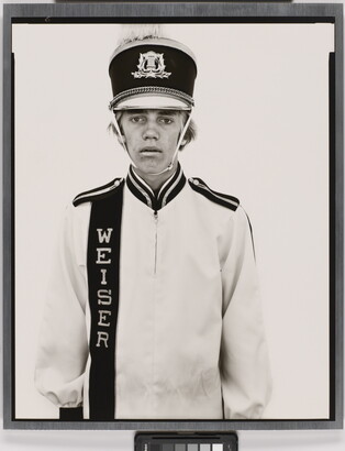 A black-and-white portrait photograph of a White teenage boy wearing an oversized, white, marching-band hat and uniform.