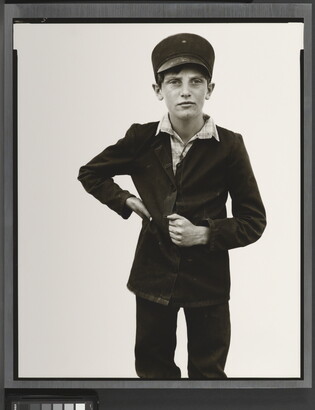 A black-and-white portrait photograph of a White boy wearing a Breton cap, a dark button-up jacket with one hand on his waist and the other tucked in between the buttons.