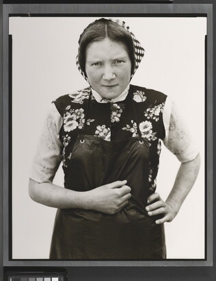 A black-and-white portrait photograph of young White woman wearing a checked kerchief, flowered vest, and dark apron.