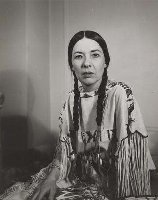 A black-and-white photograph of a seated Indigenous woman, hair in braids, gazing into the camera. 