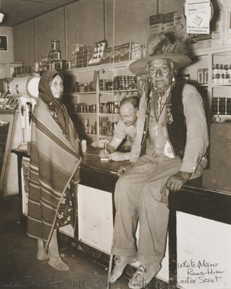 A sepia-toned photograph of a Indigenous man sitting on a counter, an Indigenous woman wrapped in a blanket standing at the counter, and a White man behind the counter.