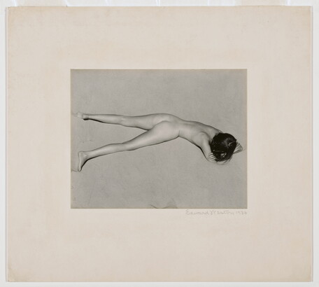 A black-and-white photograph of a nude, fair-skinned woman lying facedown in the sand with her head resting on her crossed arms.