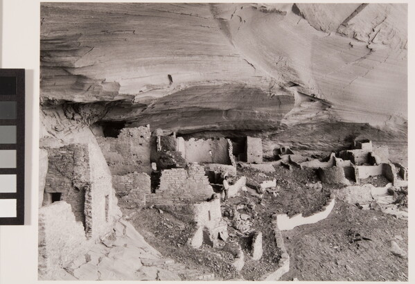 A black-and-white photograph of adobe ruins under an overhanging cliff ledge.