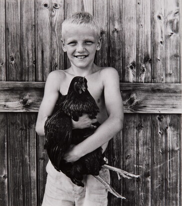 A black-and-white photograph of a smiling, shirtless White boy holding a chicken to his chest.