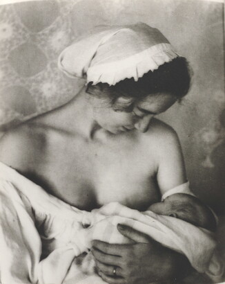 A black-and-white photograph of a White woman in a nightcap and white flowing blouse nursing an infant.